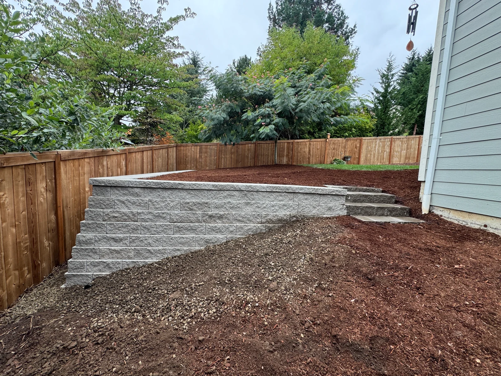 Gray segmental retaining wall with stone steps, mulch, and mature landscaping in residential backyard