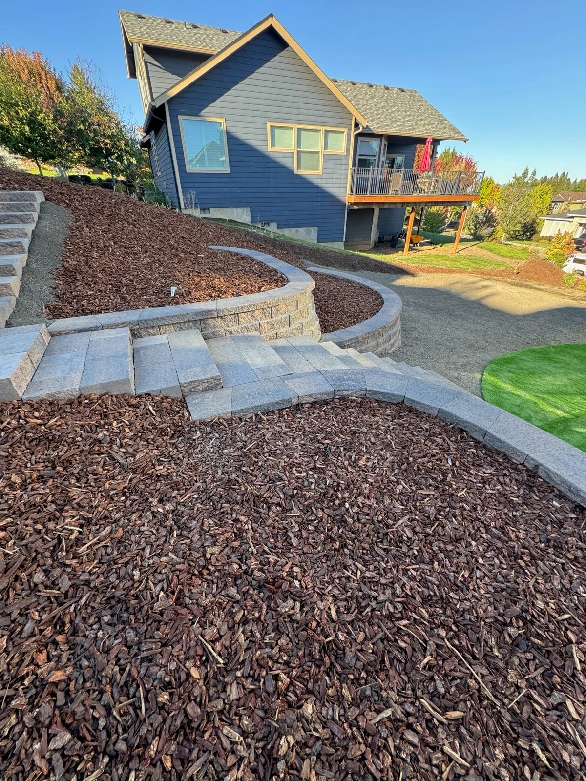 Curved stone retaining wall with concrete paver steps and dark mulch landscaping on sloped yard