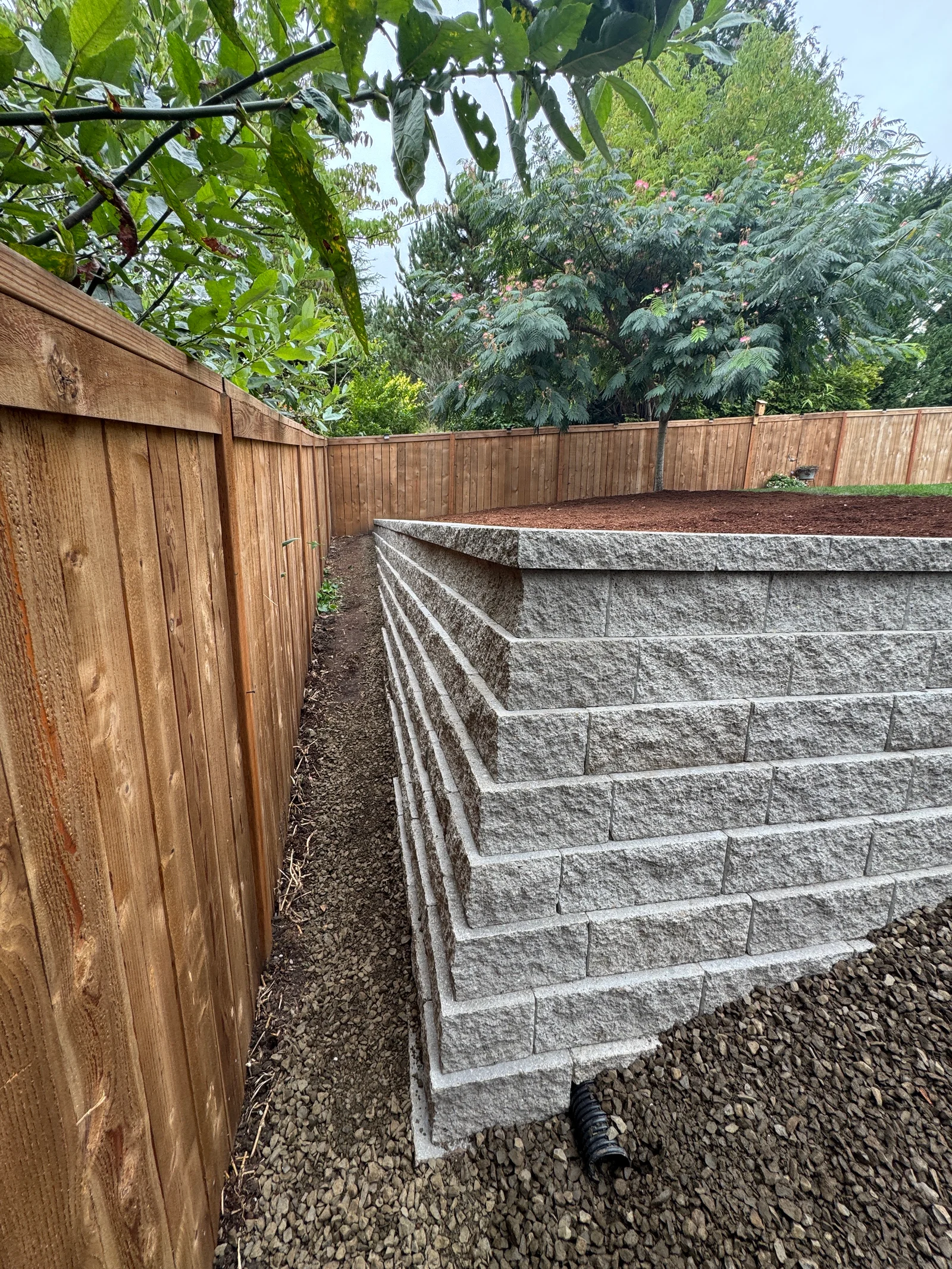 Gray concrete retaining wall with multiple tiers, wood privacy fence, and gravel base in residential backyard