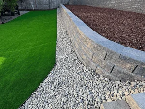 Artificial turf meets stone-edged mulch bed with gravel base and retaining wall