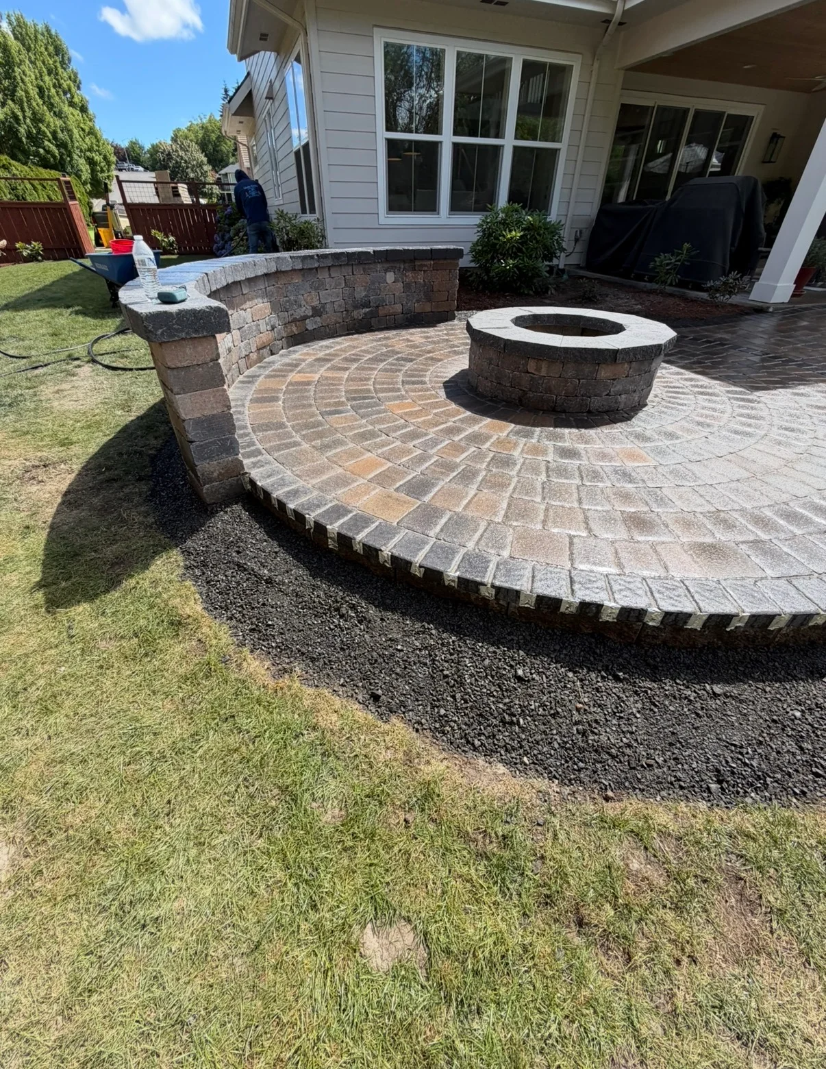 Circular paver patio with radial pattern, central fire pit, curved retaining wall, and stone pillar
