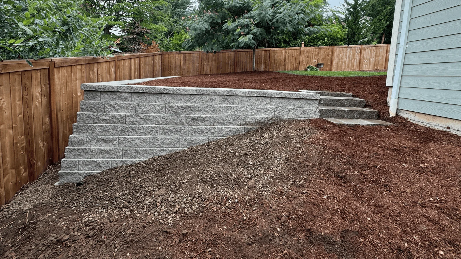 Gray segmental retaining wall with steps, gravel base, and mulched yard area