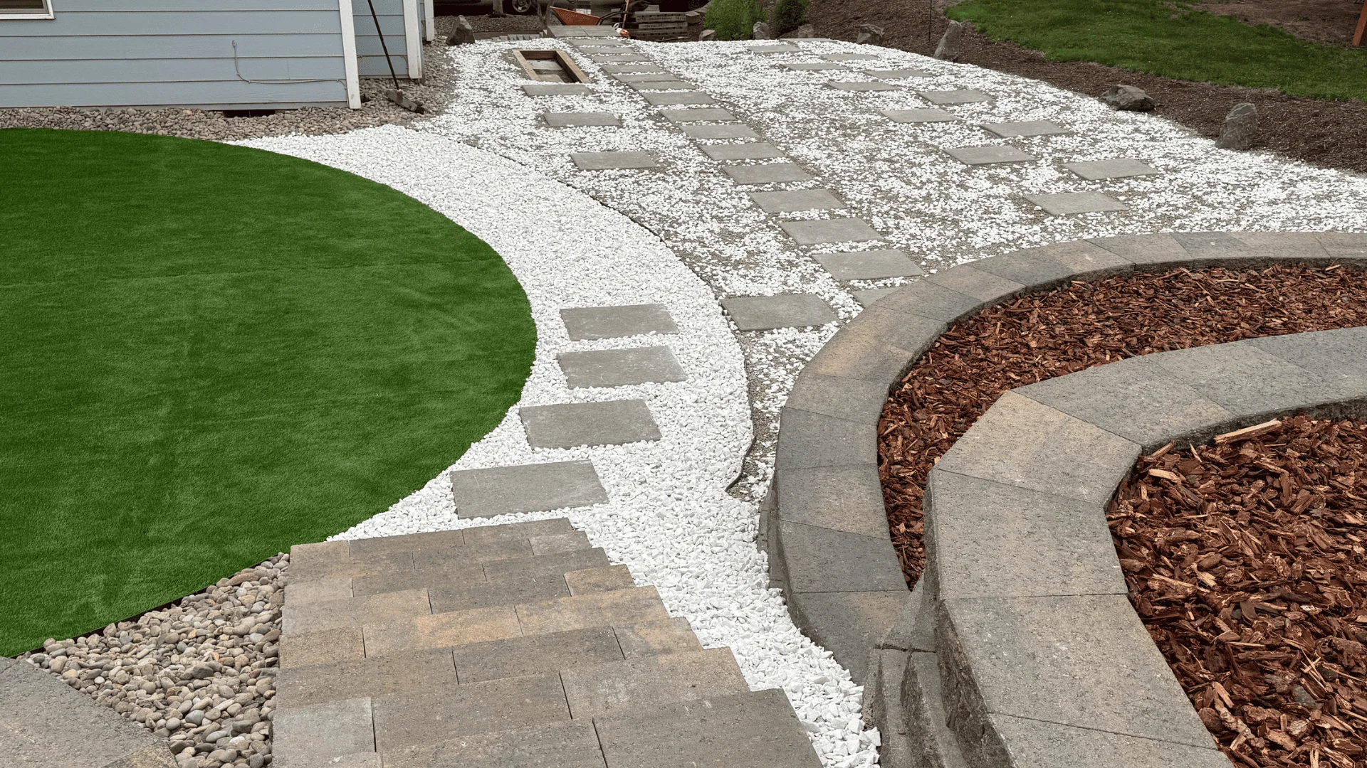 Beautiful hardscape design with synthetic grass, stepping stones, white gravel, and curved retaining wall